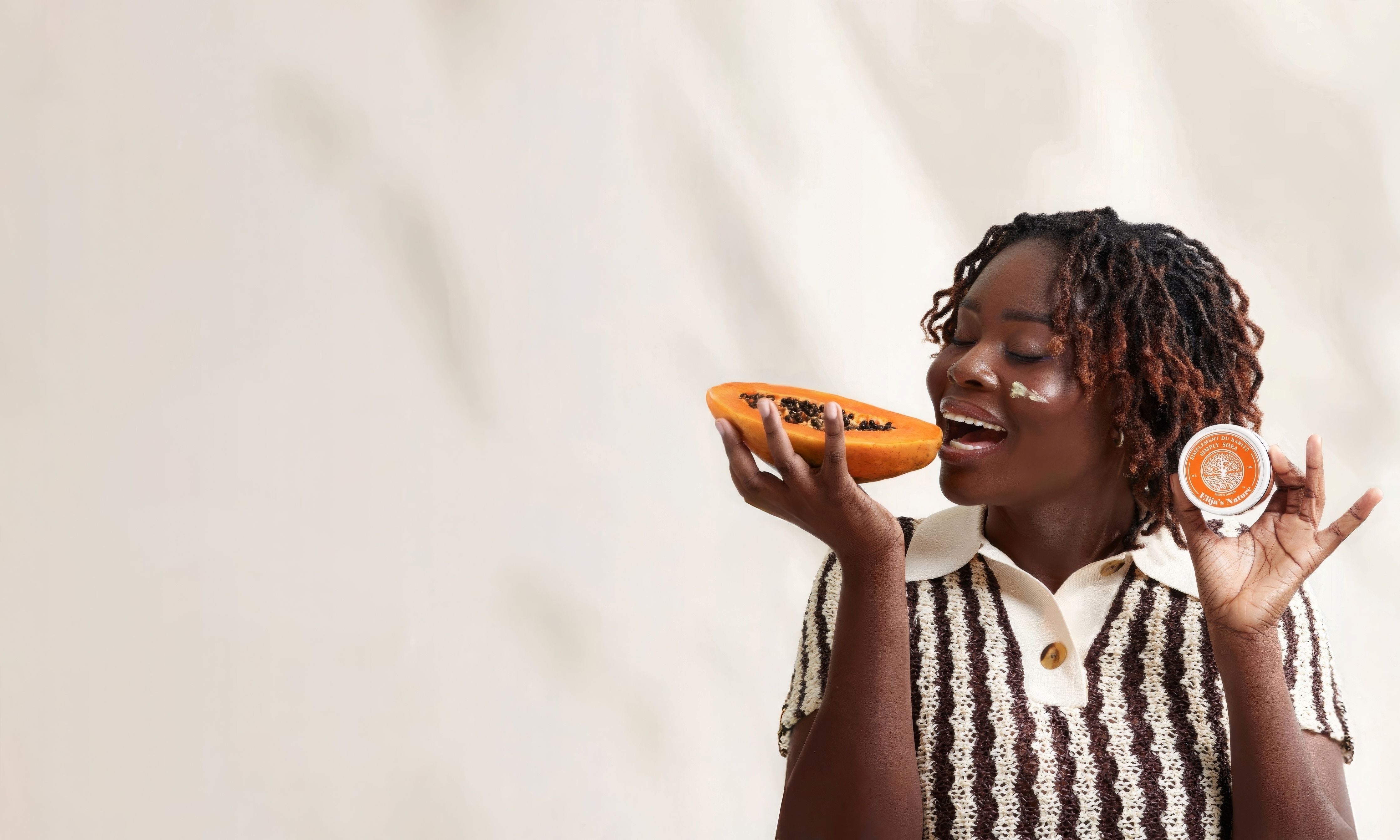 Smiling woman in striped top holding half a papaya and a small jar of Elija’s Nature product in a playful pose