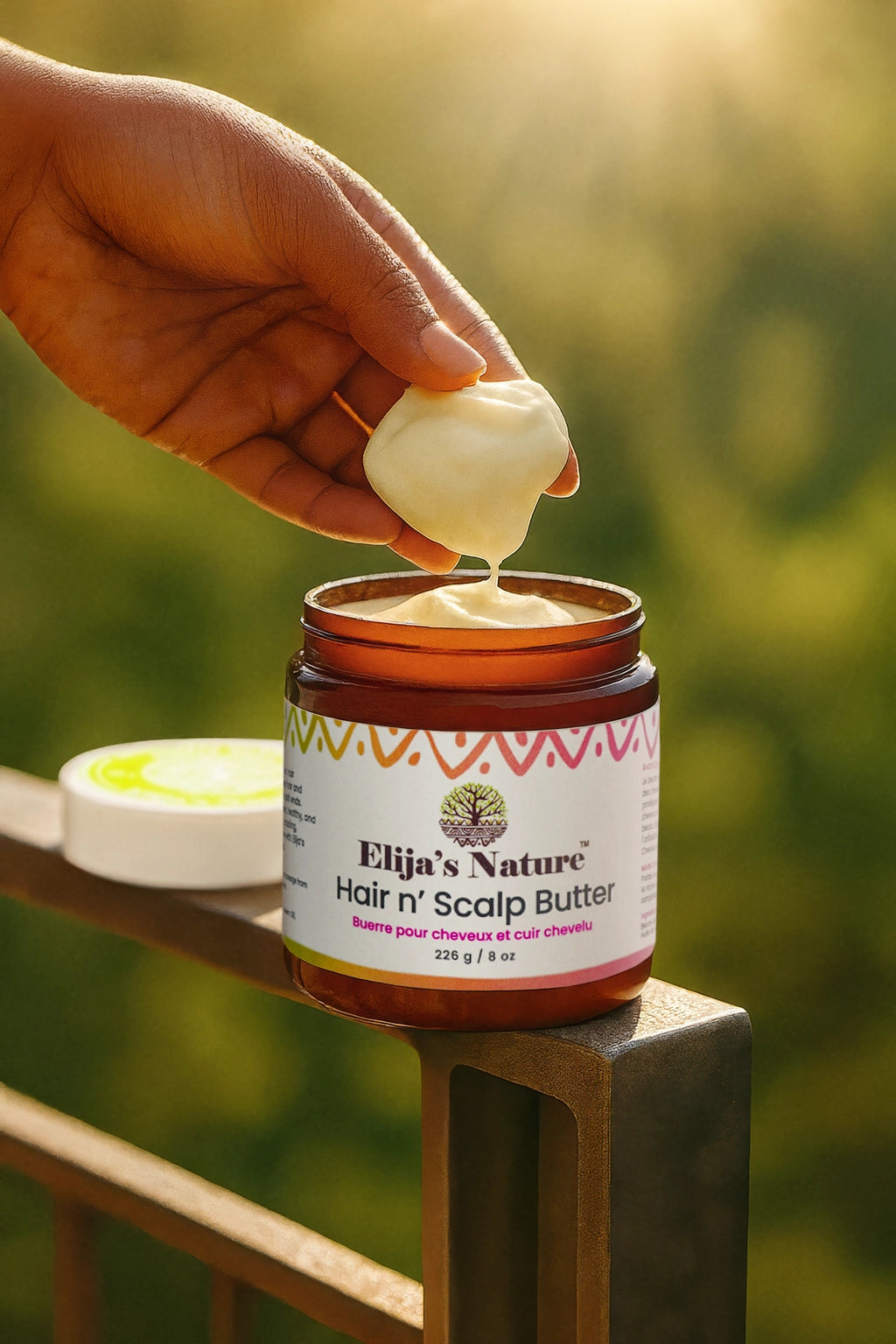 Hair N Scalp Butter - #size_8oz - Hand scooping Elija’s Nature Hair n’ Scalp Butter from an open jar placed on a metal railing in sunlight