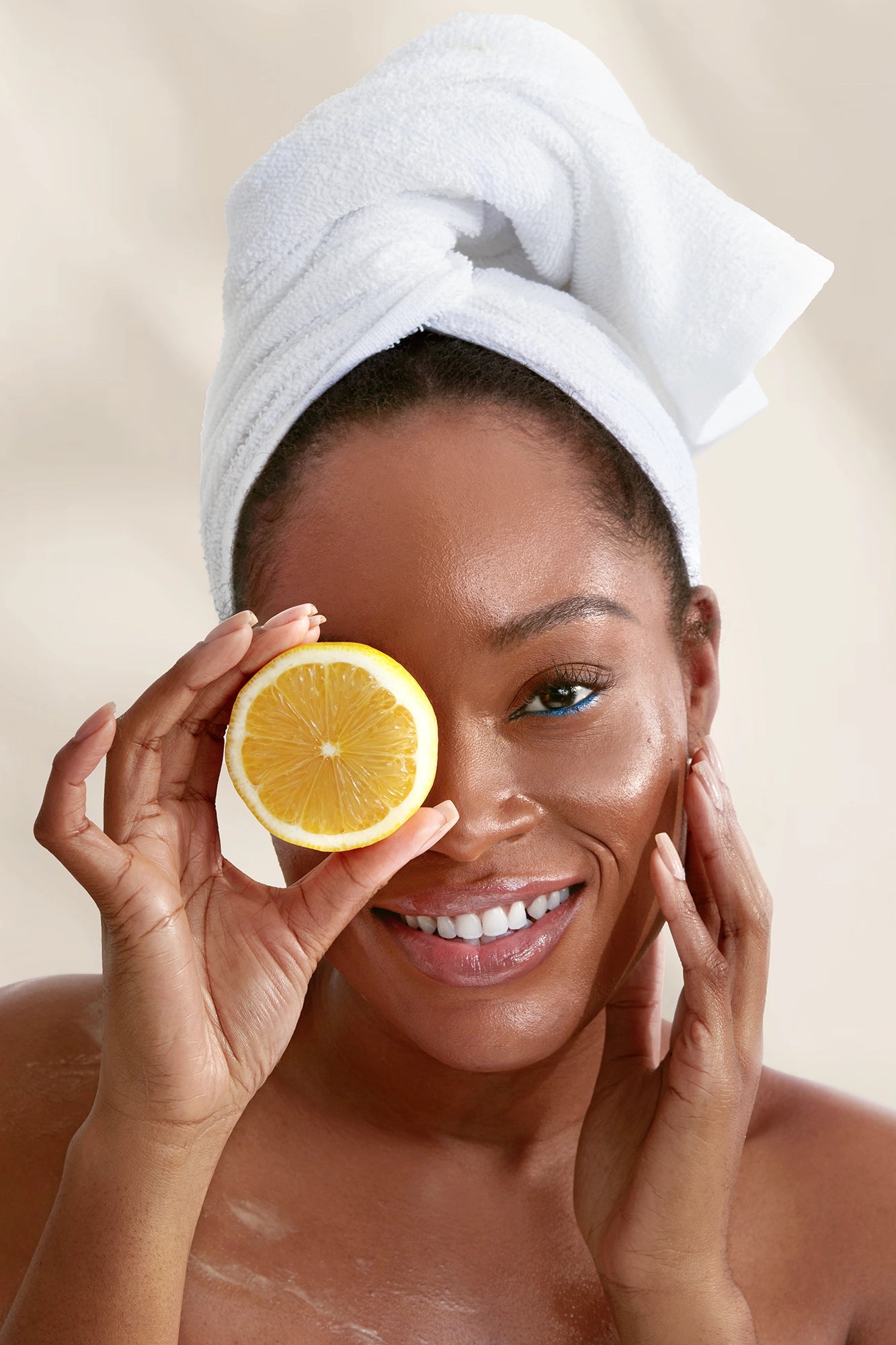 Smiling woman with a towel wrapped around her head holding a lemon slice over one eye, posing against a soft beige background