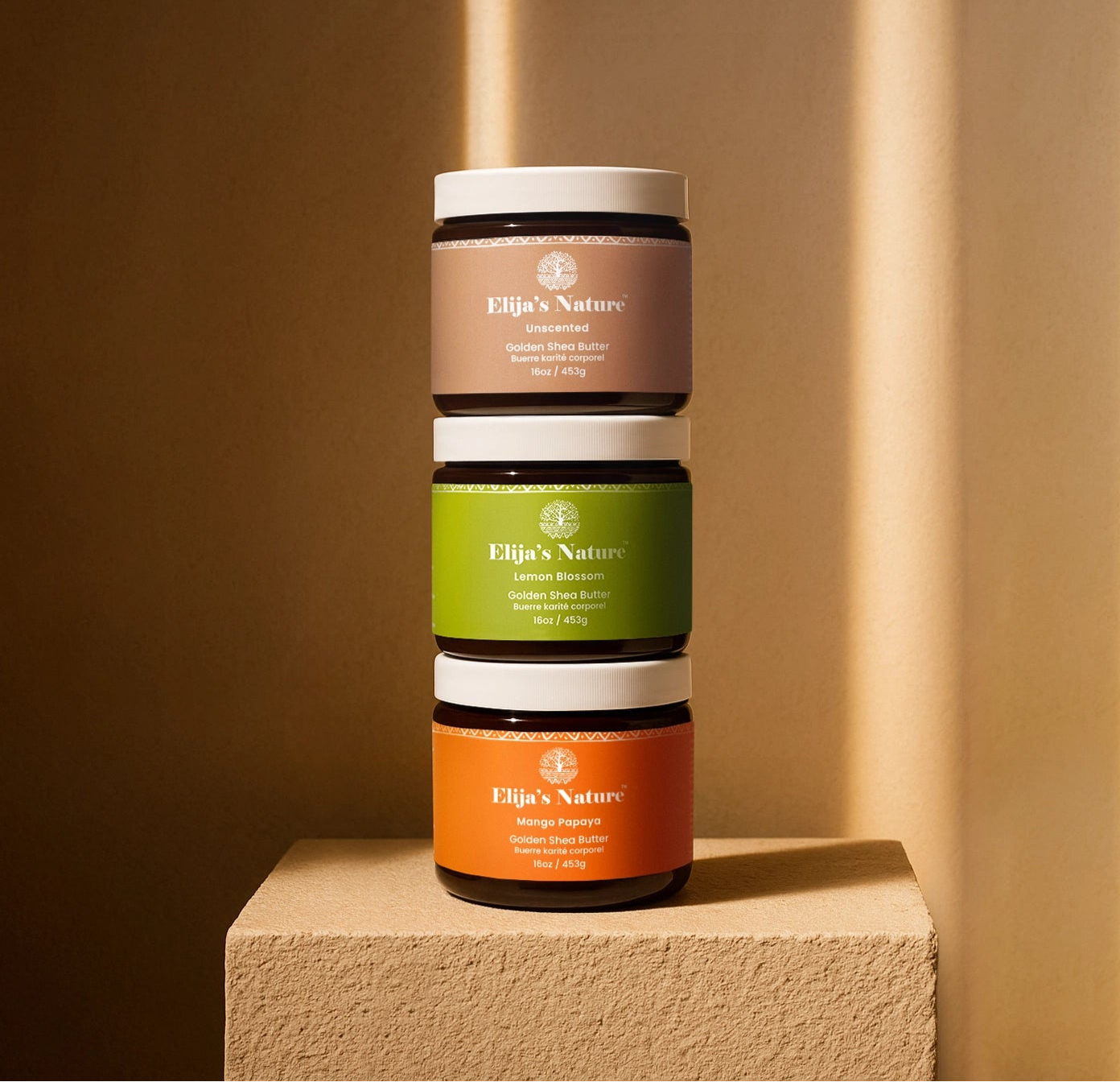Stacked trio of Elija’s Nature body butter jars: Unscented, Lemon Blossom, and Mango Papaya on a pedestal against a beige-toned wall