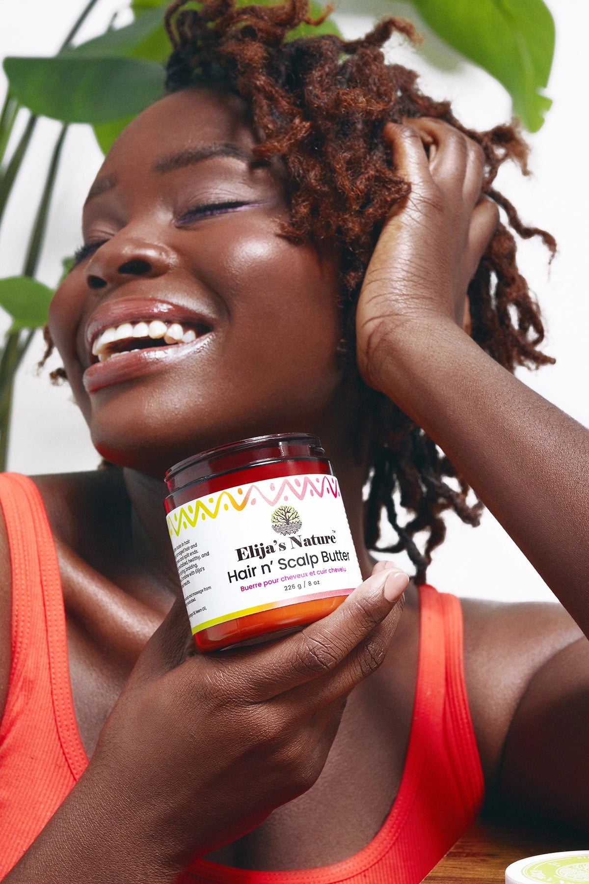 Hair N Scalp Butter - #size_8oz - Smiling woman with textured hair holding Elija’s Nature Hair n’ Scalp Butter jar beside her face, surrounded by greenery