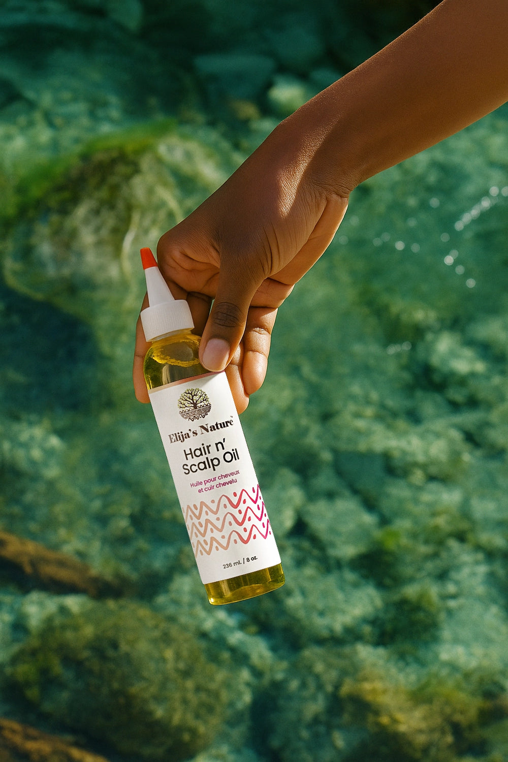 Hair N' Scalp Oil - #size_8oz - Hand holding Elija’s Nature Hair & Scalp Oil bottle over clear turquoise water in an outdoor setting