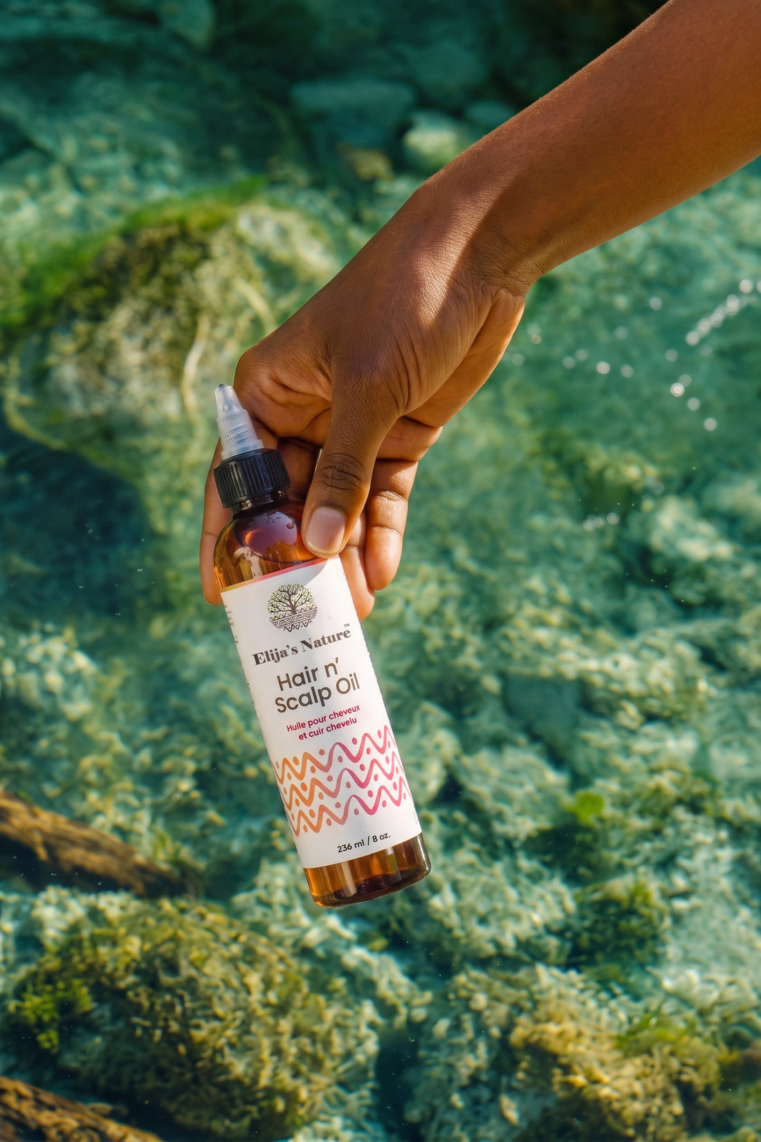 Hair N' Scalp Oil - #size_8oz - Hand holding Elija’s Nature Hair & Scalp Oil bottle over clear turquoise water in an outdoor setting