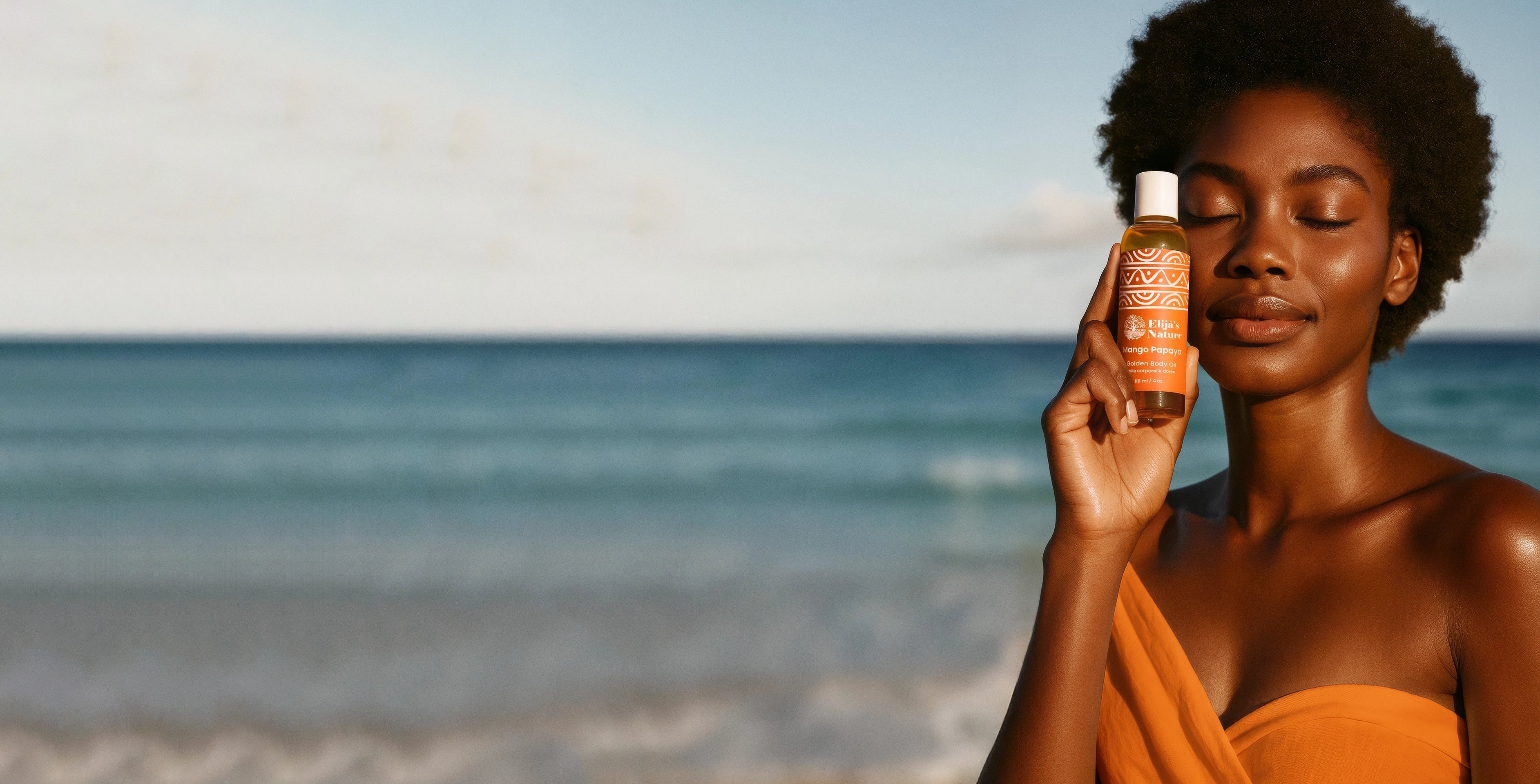Woman holding Elija’s Nature's Mango Papaya Body Oil beside her face, smiling with closed eyes at the beach