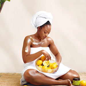 Golden Body Butter - #scent_Lemon Blossom #size_16oz -  A woman seated cross-legged in a towel applies body butter while holding a bowl of fresh lemons, conveying freshness and citrus-infused skincare
