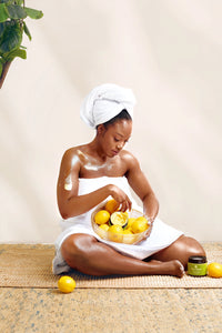 Golden Body Butter - #scent_Lemon Blossom #size_16oz -  A woman seated cross-legged in a towel applies body butter while holding a bowl of fresh lemons, conveying freshness and citrus-infused skincare