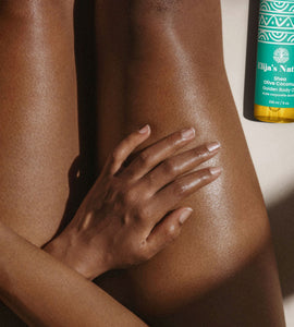 #scent_Olive Coconut #size_8oz - Woman with glowing skin sitting next to an Elija's Nature Golden Beauty Oil in the scent Olive Coconut